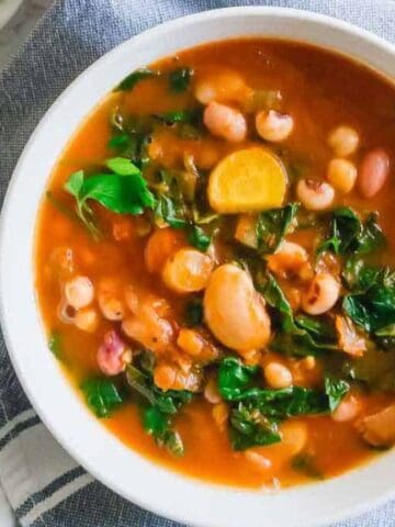 A bowl of vegetable and bean soup with leafy greens, placed on a blue cloth next to a bowl of chopped herbs and gold utensils.