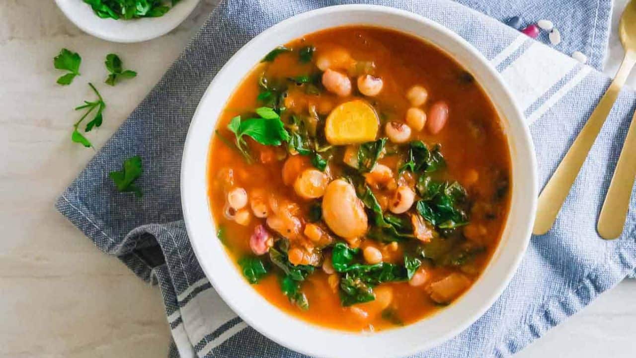 A bowl of vegetable and bean soup with leafy greens, placed on a blue cloth next to a bowl of chopped herbs and gold utensils.