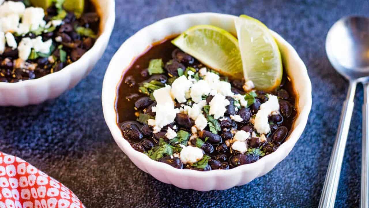 Low angle shot of two bowls of Mexican black beans garnished with crumbled cheese and lime wedges.