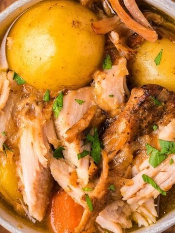 A bowl of shredded pork roast with potatoes, carrots, and chopped parsley in broth, viewed from above on a wooden surface.