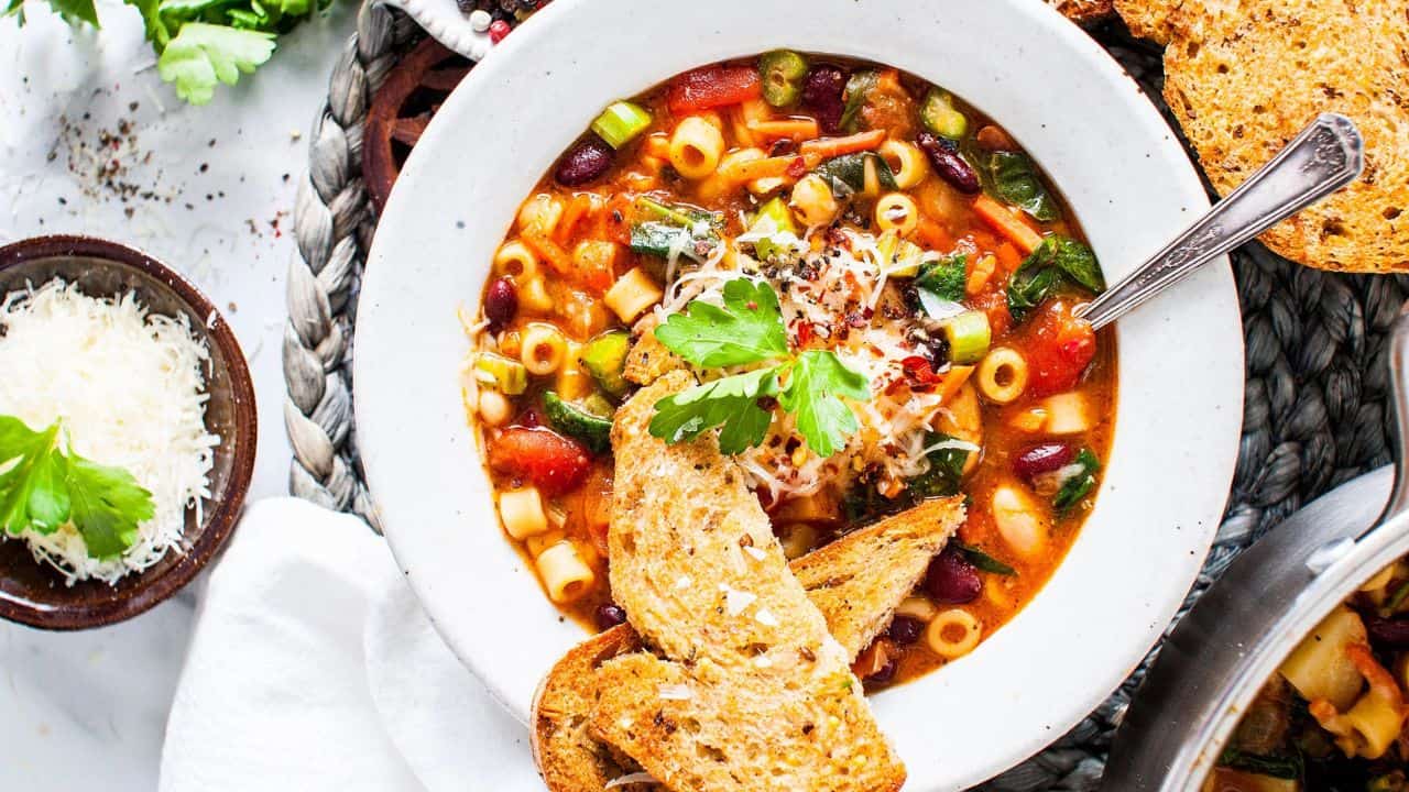 A bowl of vegetable soup with pasta, beans, greens, and tomato broth, topped with grated cheese and parsley, served with toasted bread slices.