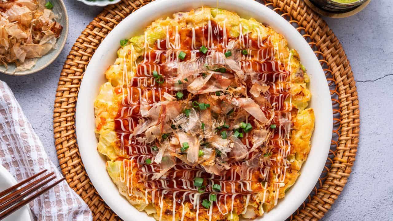 A plate of okonomiyaki topped with mayonnaise, okonomiyaki sauce, bonito flakes, and chopped green onions on a woven placemat.