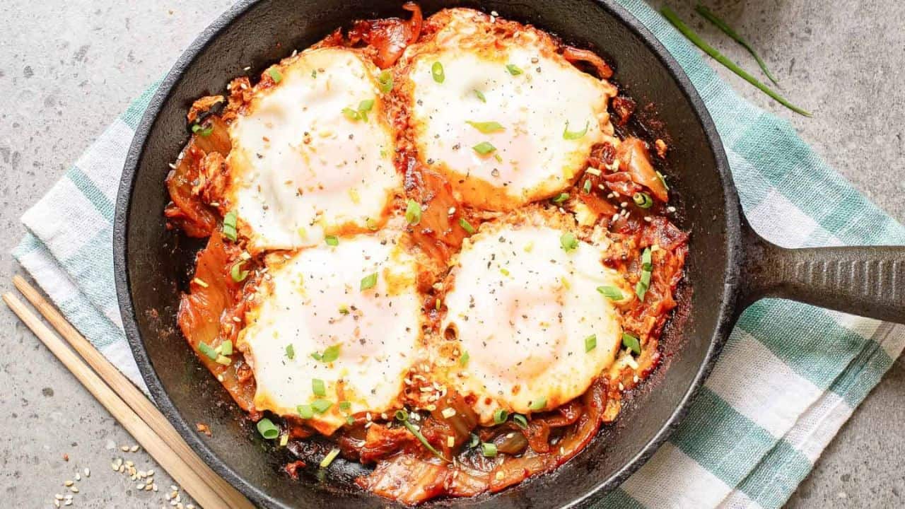 A hand dips bread into a skillet of shakshuka, featuring poached eggs, tomato sauce, and garnished with chopped green onions.