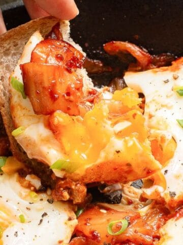 A hand dips a piece of bread into a skillet with baked eggs in tomato sauce, garnished with green onions.