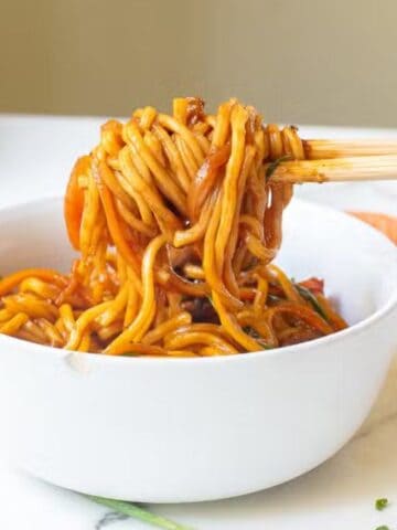 A hand uses chopsticks to lift saucy noodles from a white bowl on a white countertop, with a carrot and a pan visible in the background.
