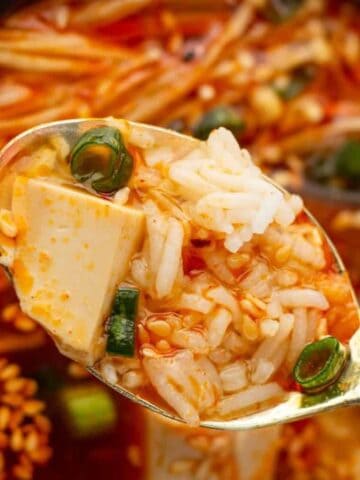 A close-up of a spoonful of spicy soup with tofu, rice, green onions, and sesame seeds over a bowl of similar soup.