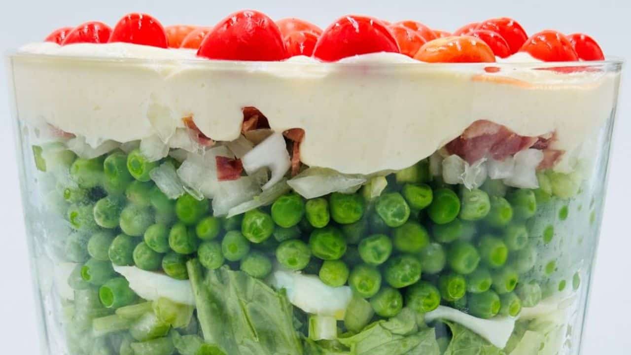 A layered salad in a clear glass trifle bowl on a wooden board, with peas, lettuce, and red tomatoes, next to a floral patterned cloth.