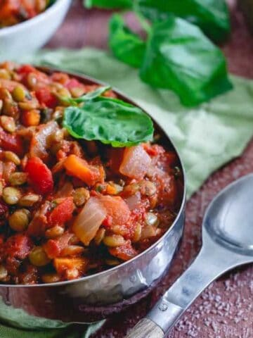 A metal bowl filled with lentil stew, garnished with a basil leaf, sits on a wooden table next to a spoon and green napkin, with more food and cheese in the background.