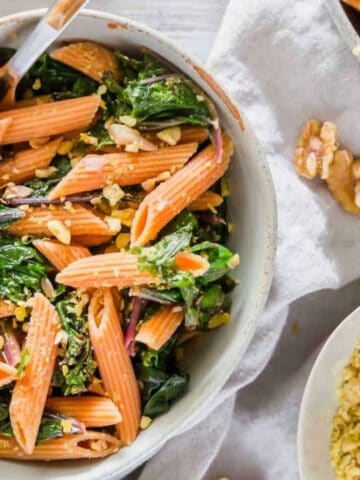 A bowl of penne pasta mixed with leafy greens and topped with crushed nuts, with additional nuts and a bowl of grated cheese nearby.