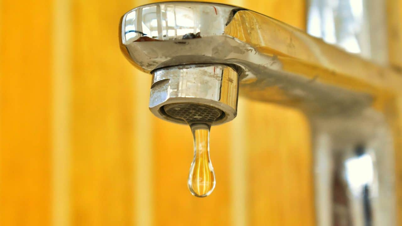 A close-up of a metal faucet with a single water droplet hanging from its spout, against a yellow striped background.