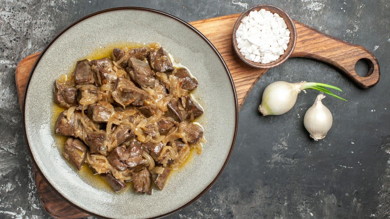 A plate of cooked liver and onions sits on a wooden board, next to a bowl of coarse salt and two whole onions on a dark stone surface.