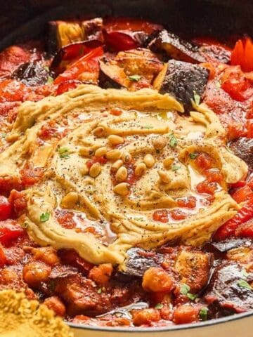 A skillet filled with a stew of tomatoes, chickpeas, and eggplant, topped with a swirl of hummus and sprinkled with pine nuts.