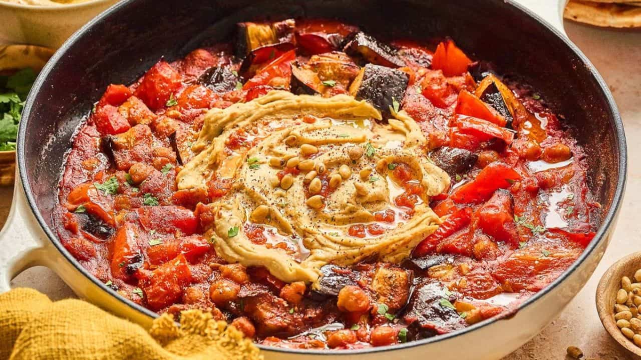 A skillet filled with a stew of tomatoes, chickpeas, and eggplant, topped with a swirl of hummus and sprinkled with pine nuts.
