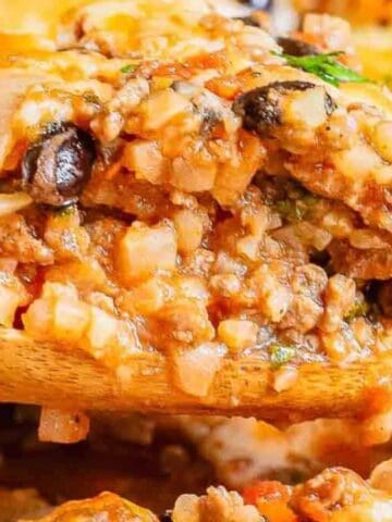 A wooden spoon scoops a serving of cheesy ground meat casserole with rice, black beans, onions, and herbs.
