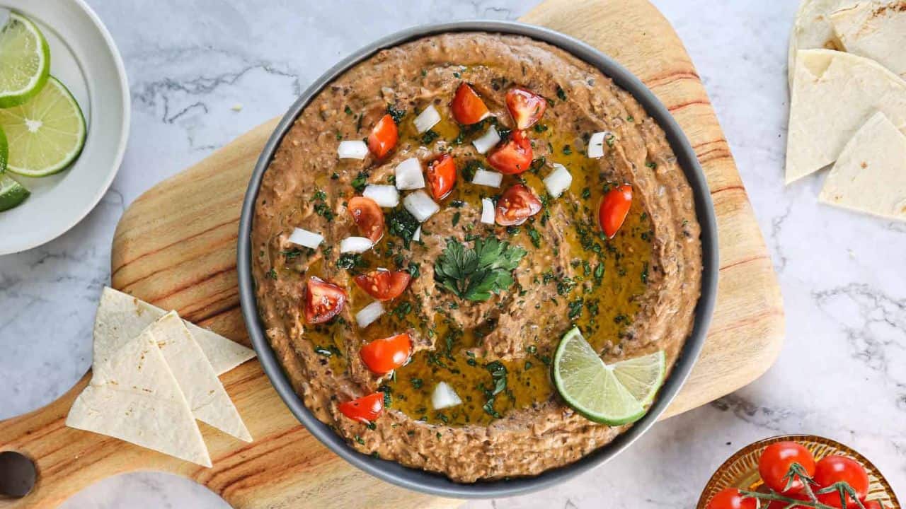 A bowl of refried beans topped with chopped tomatoes, diced onions, fresh parsley, and a lime wedge sits on a wooden board. Tortilla pieces and lime slices are visible nearby.