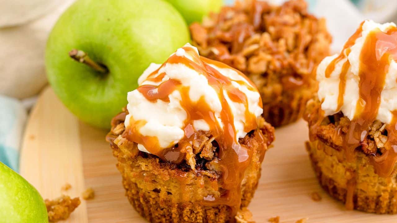 Close-up of two apple crumble cupcakes topped with whipped cream and caramel drizzle, placed on a wooden surface with green apples in the background.
