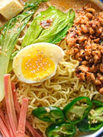 A bowl of ramen topped with half a boiled egg, tofu, greens, sliced meat, jalapeños, minced meat, and chopsticks resting on the bowl's edge.