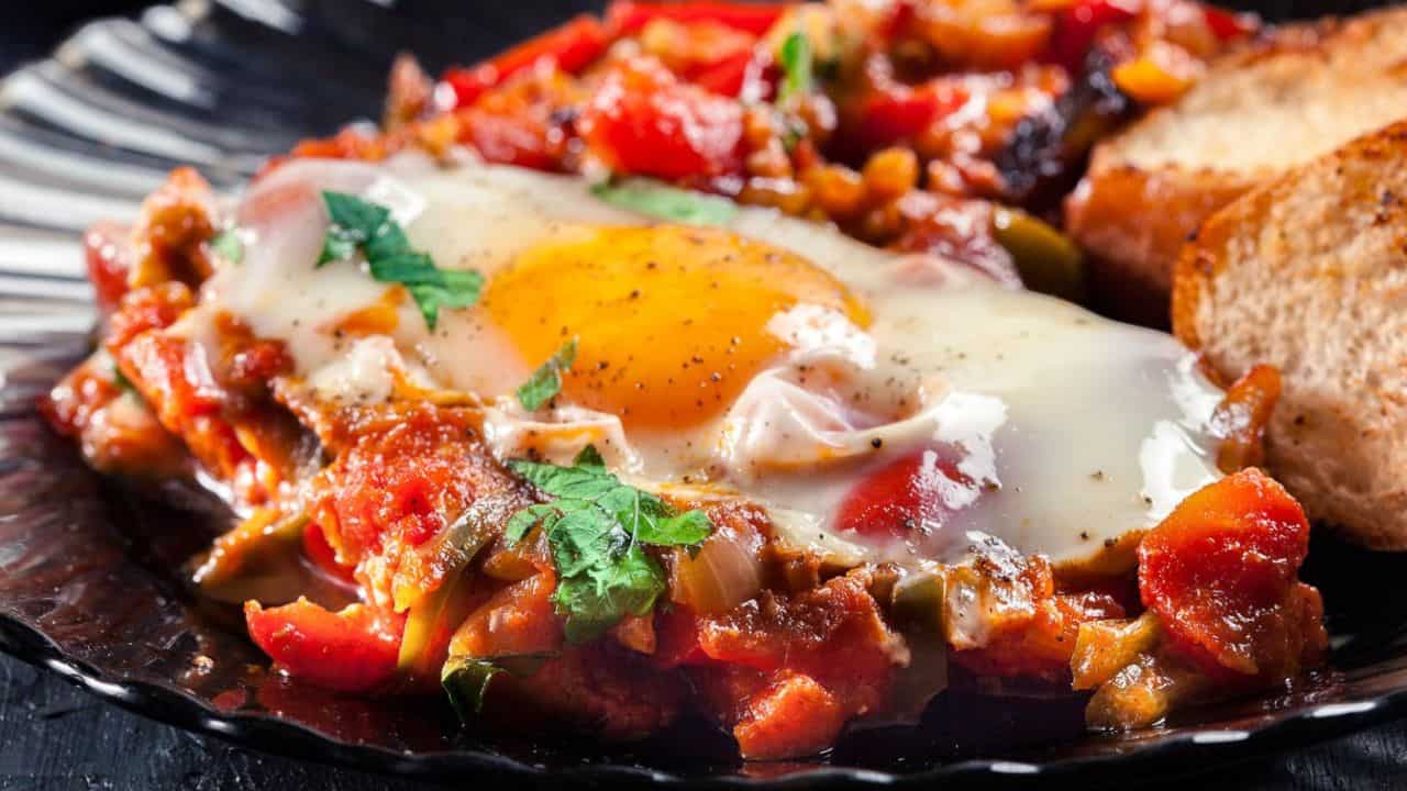 A plate with Moroccan Shakshuka, two slices of toasted bread, and chopped herbs, with a pan of shakshuka, tomatoes, and a bowl of greens in the background—a delicious taste of North African cuisine.