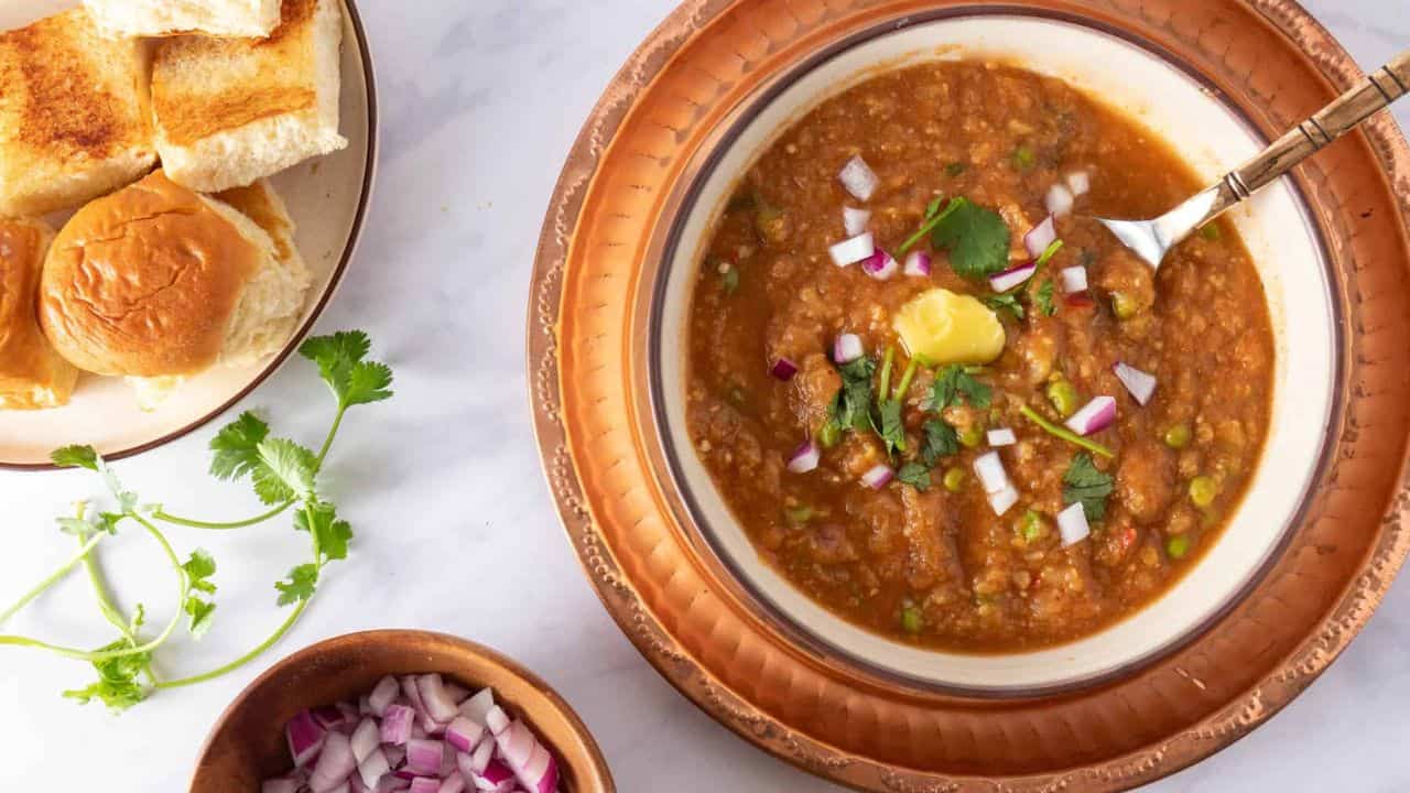 A top-down view of a plate with pav rolls, bhaji masala, red onions, lemon wedges, and garnishes on the side.