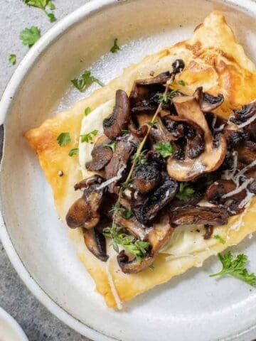 A square puff pastry topped with sautéed mushrooms and herbs is served on a round white plate placed on a gray surface.