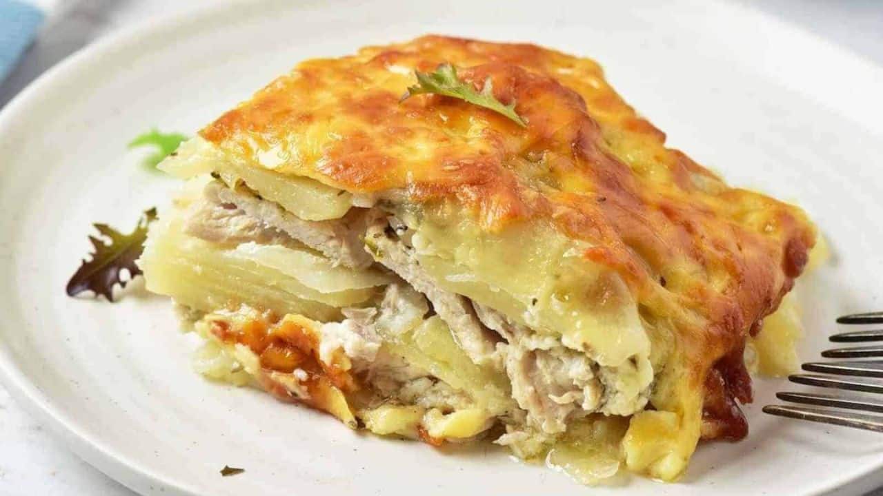 A slice of cheesy potato casserole with layers of potatoes, shredded meat, and melted cheese served on a white plate with a fork and a small salad garnish.