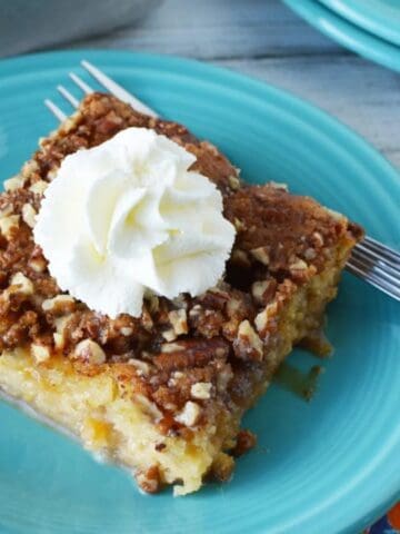 A square piece of cake topped with chopped nuts and a swirl of whipped cream sits on a turquoise plate with a fork.