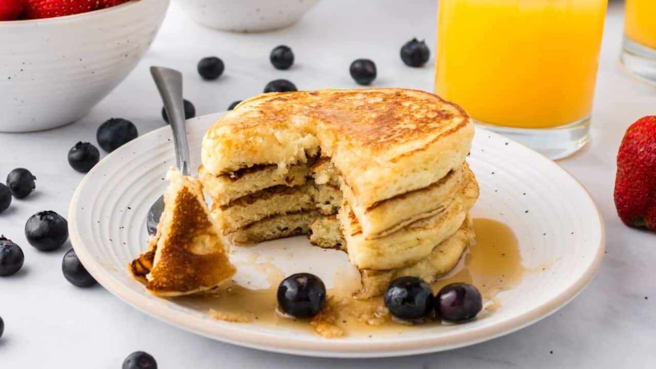 A stack of pancakes with syrup and blueberries on a white plate, a fork taking a piece, with a glass of orange juice and a bowl of strawberries in the background.