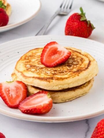Two pancakes topped with sliced strawberries on a white plate, with whole strawberries, a fork, and a glass of orange juice nearby.