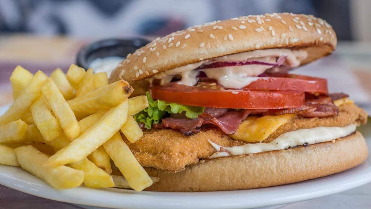 A plate with French fries and a breaded chicken sandwich topped with lettuce, tomato, bacon, cheese, red onion, mayonnaise, and sauce in a sesame seed bun.