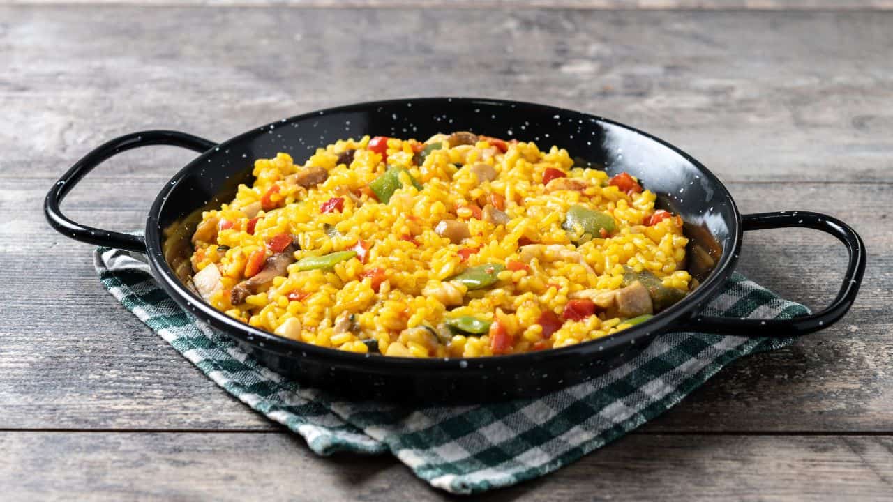A round black pan filled with yellow rice, vegetables, and pieces of meat sits on a green and white checkered cloth on a wooden table.