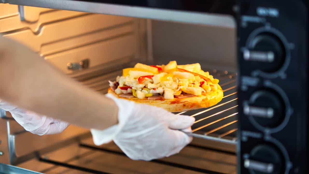 A person wearing white gloves places an uncooked pizza with vegetables and pineapple onto the rack inside an oven.