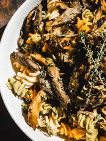 A bowl of tri-color rotini pasta topped with sautéed mushrooms and garnished with fresh herbs, placed on a wooden surface next to a fork and knife.