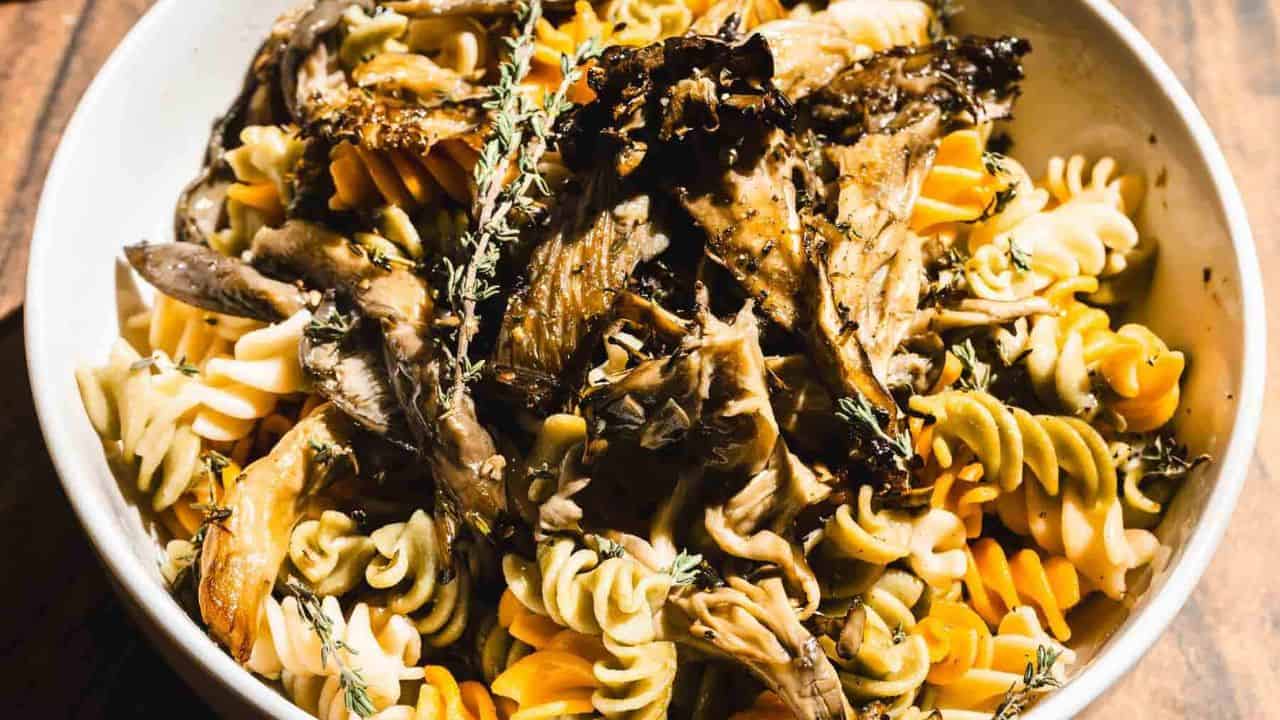 A bowl of multicolored fusilli pasta topped with cooked mushrooms and garnished with fresh thyme sprigs. The dish is served in a white bowl on a wooden surface.