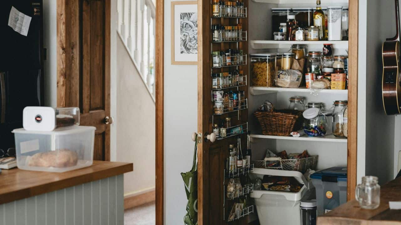 A kitchen pantry with open shelves and a door, stocked with jars, cans, bottles, baskets, and various food items. A countertop with a loaf of bread is visible in the foreground.