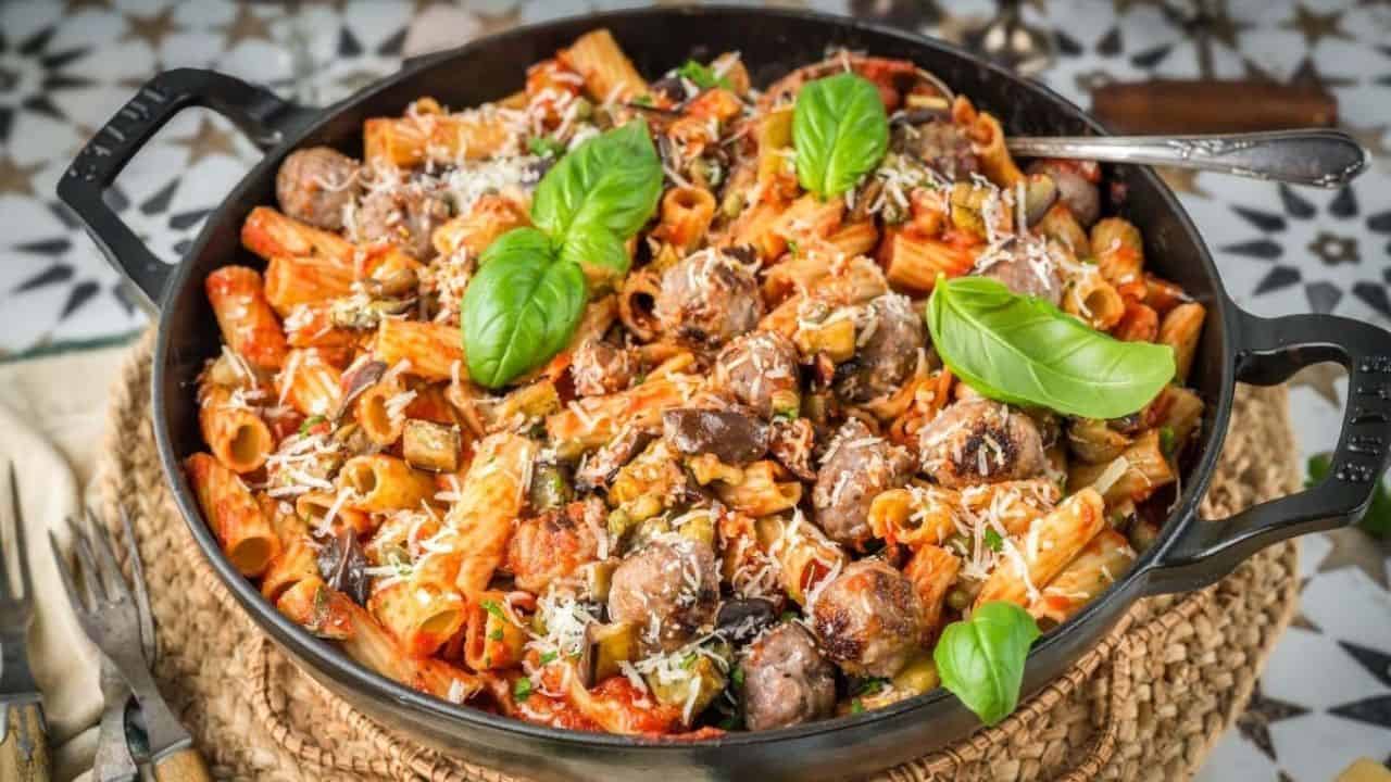 A skillet filled with rigatoni pasta, sausage, tomato sauce, mushrooms, grated cheese, and garnished with fresh basil leaves.