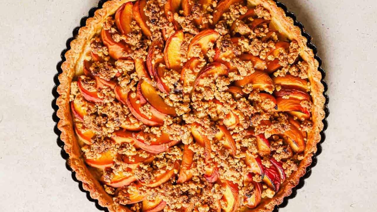 A baked peach tart with a golden brown crust, topped with peach slices and streusel crumble, on a light-colored surface.
