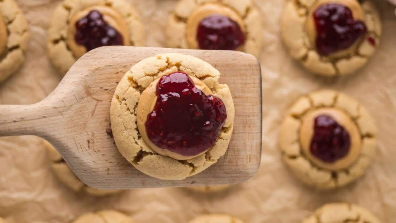 A wooden spatula holds a thumbprint cookie topped with red jam, with more cookies on parchment paper in the background.