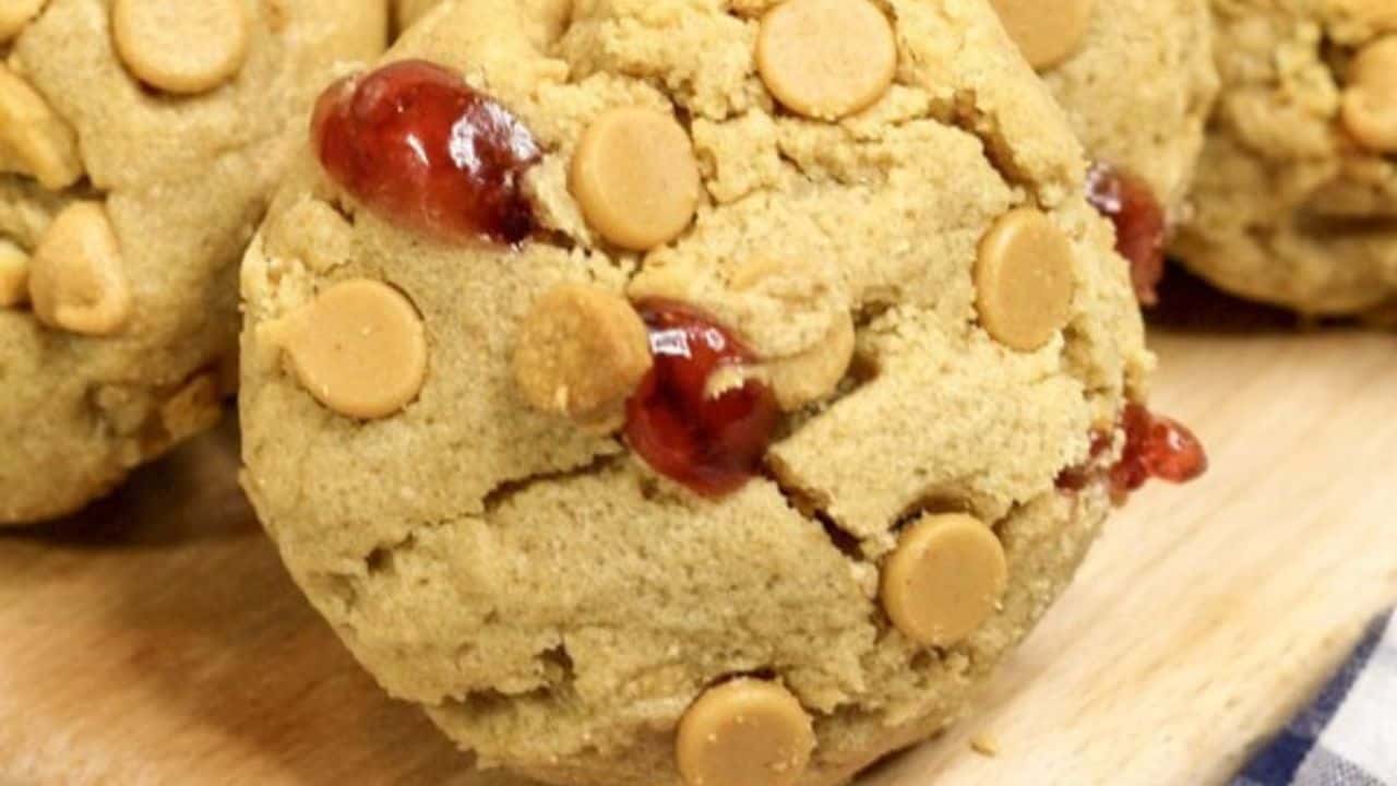 A close-up of a cookie with peanut butter chips and visible spots of red jelly on a wooden surface.