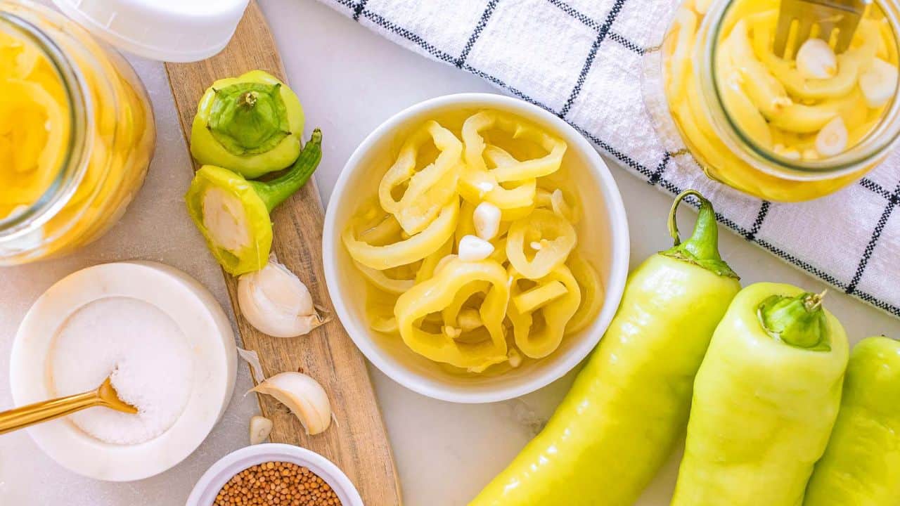 A bowl of sliced pickled banana peppers, whole banana peppers, garlic cloves, mustard seeds, salt, and jars of pickled peppers on a white surface with a checkered towel.