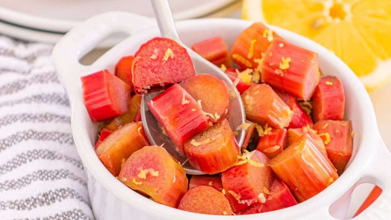 A bowl of chopped rhubarb pieces with lemon zest, and a spoon scooping some pieces. A sliced lemon is visible in the background.