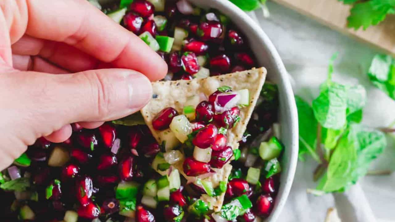 A hand holds a chip topped with pomegranate salsa, featuring diced cucumber, red onion, and herbs in a bowl.