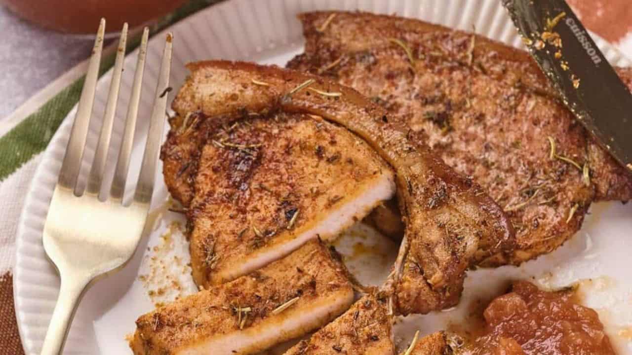 A plate with seasoned, cooked pork chops, some sliced, served with a gold fork and knife, and a portion of brown sauce or chutney on the side.