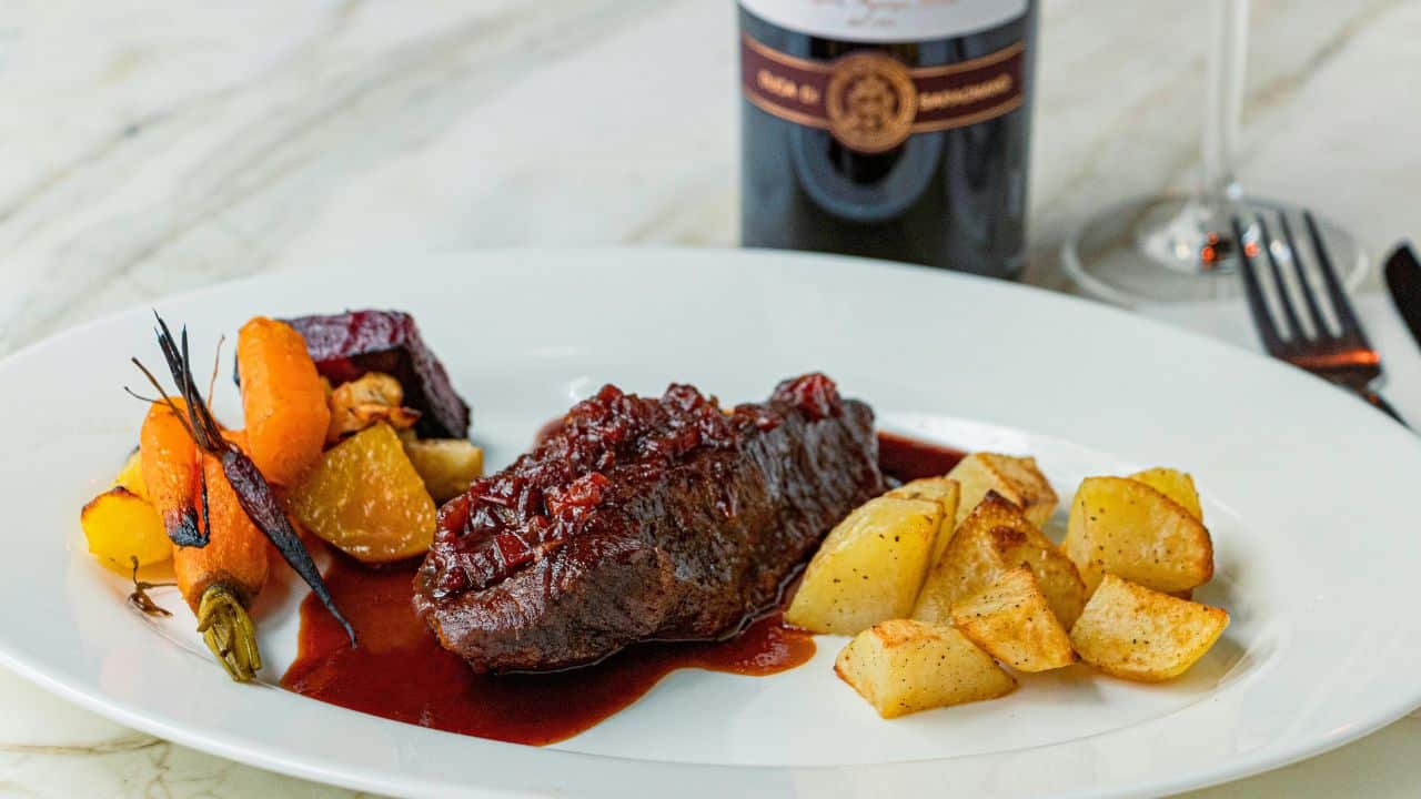 A plated meal with roasted potatoes, glazed carrots and beets, and a piece of meat topped with a red sauce, next to a wine bottle and glass on a marble surface.