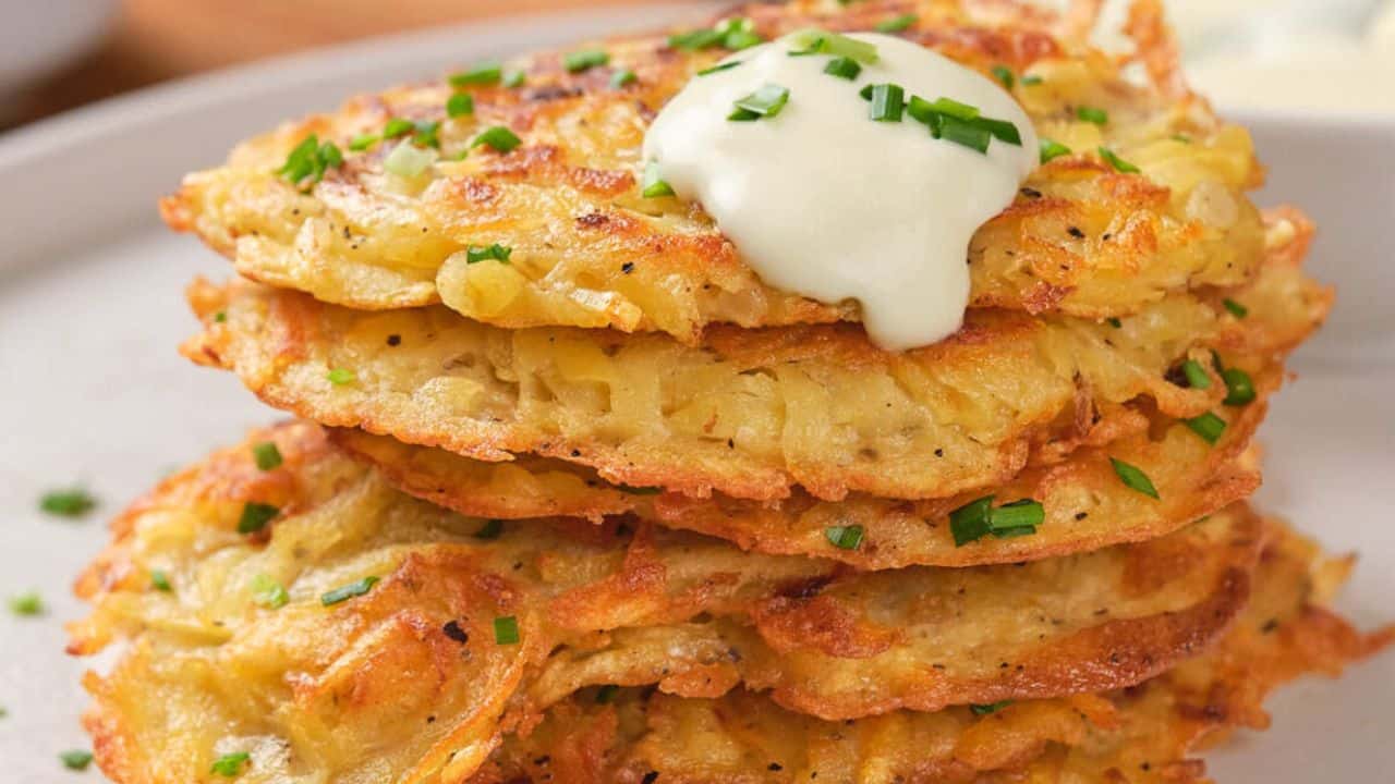 A stack of crispy potato pancakes garnished with chopped chives and topped with a dollop of sour cream on a white plate.