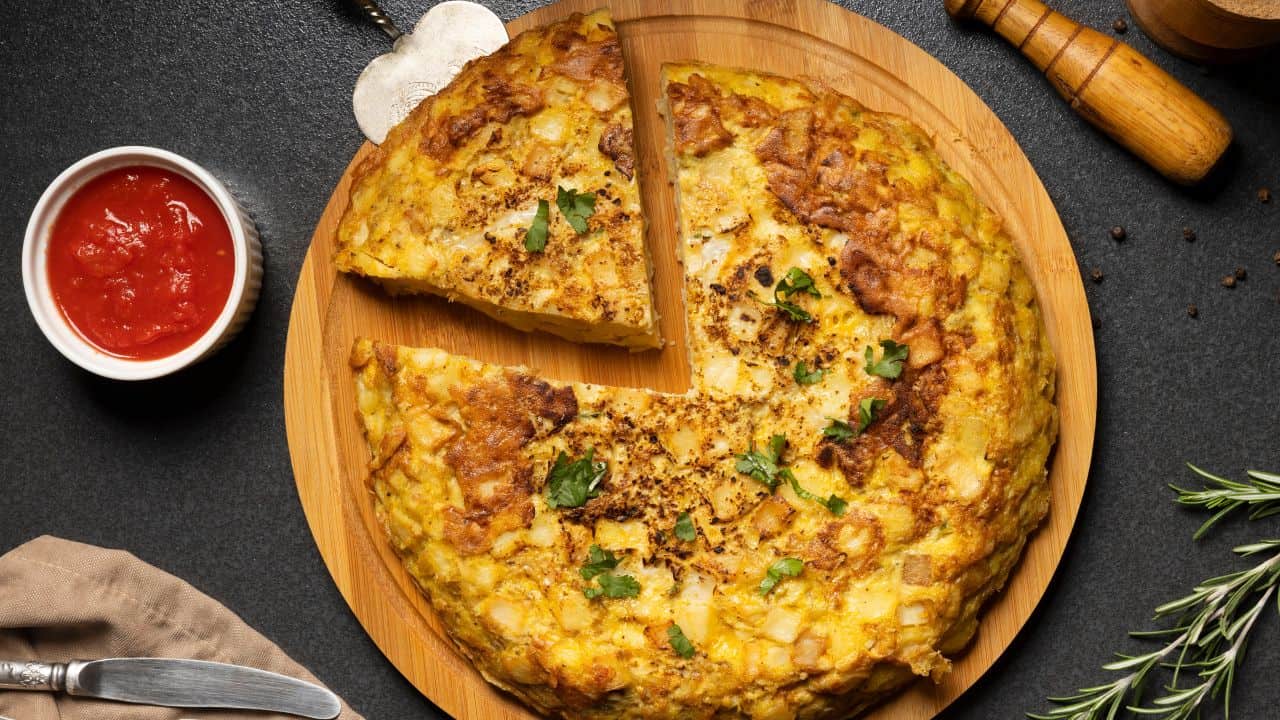 A Spanish omelette with a slice cut out sits on a wooden plate, garnished with parsley, next to a small bowl of red sauce and a wooden pepper grinder.
