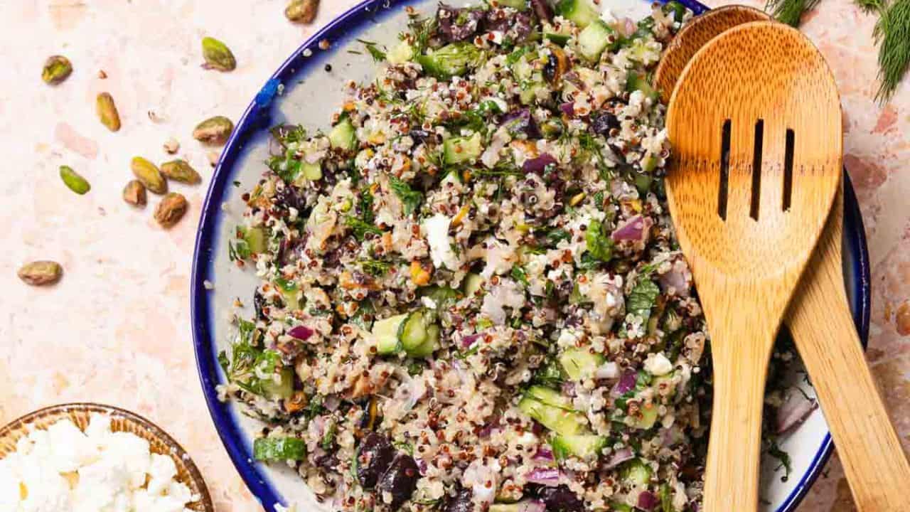 A white plate with a serving of quinoa salad mixed with diced cucumbers, chopped herbs, red onions, nuts, and feta cheese. A gold fork is resting on the plate. The background is a light, textured surface.