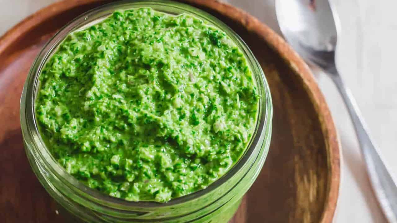 Radish greens pesto in a glass jar on a wooden plate with a serving spoon.
