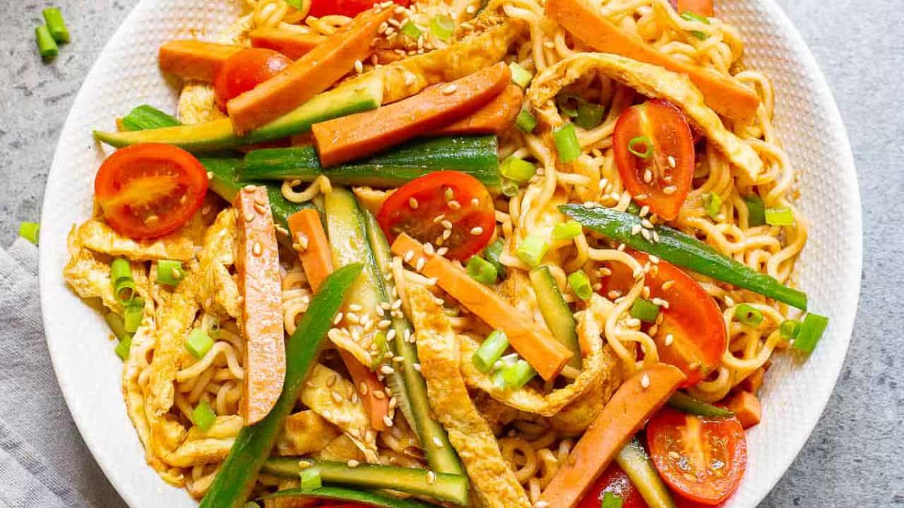 A bowl of stir-fried noodles with chopsticks lifting a portion. The dish includes sliced cucumbers, tomatoes, and green onions. A sauce jug and small bowl are in the background. The scene is set on a stone countertop.