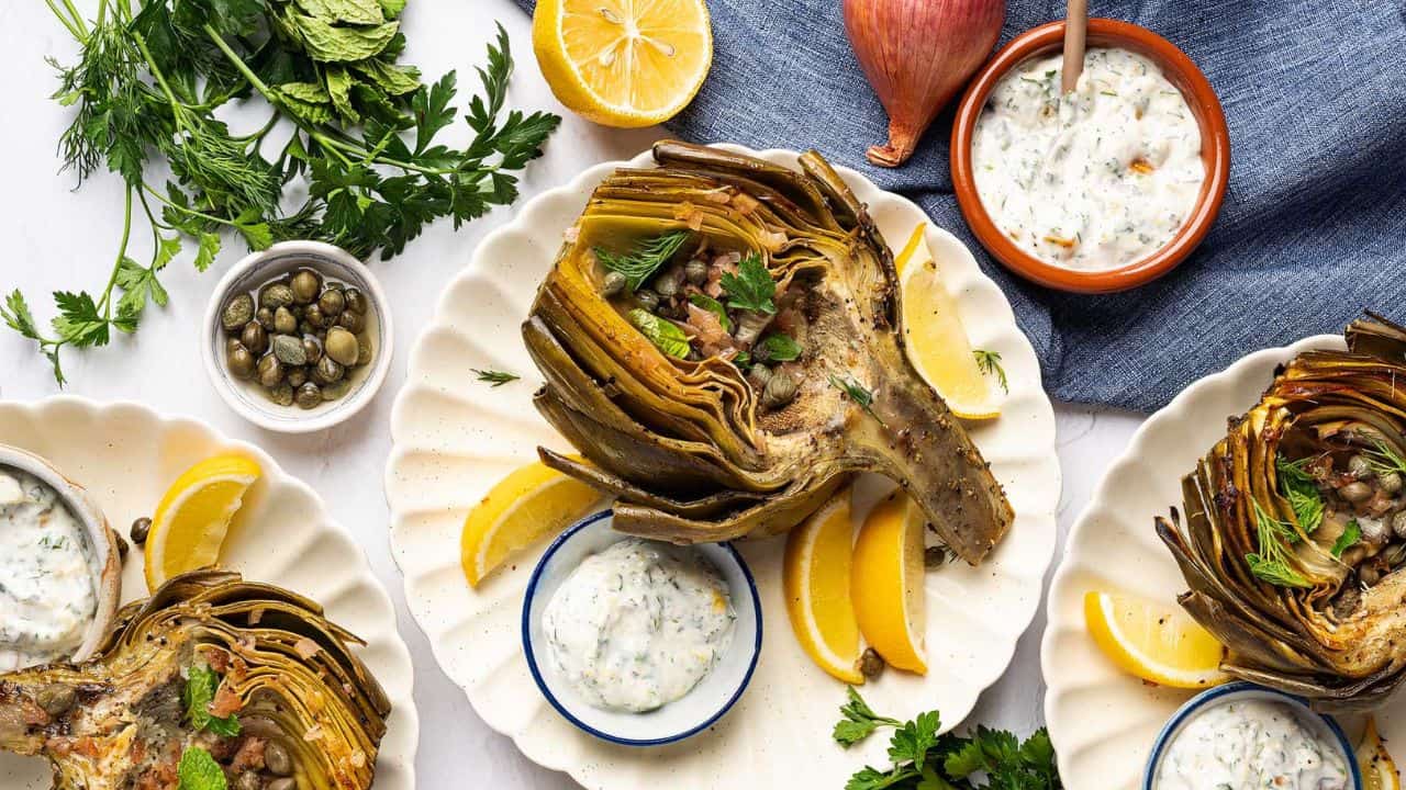 Roasted artichoke halves on plates with lemon wedges, creamy herb dip, fresh herbs, and capers, arranged on a white surface with a blue cloth and a shallot.