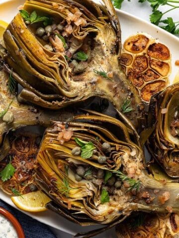 Roasted artichoke halves garnished with herbs and capers, served with lemon wedges, garlic, a glass of white wine, sauce, and fresh parsley on a white surface.
